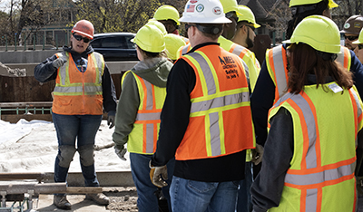 A woman in a yellow safety vest speaks to a crowd of trainees, who are also wearing safety vests.